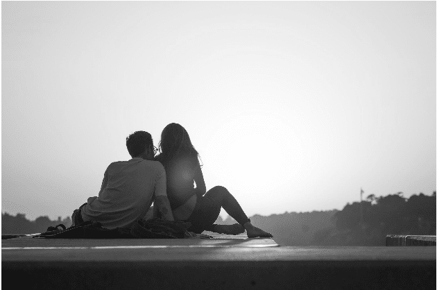 A couple sitting on a ledge looks at the sunset, with trees visible in the distance.