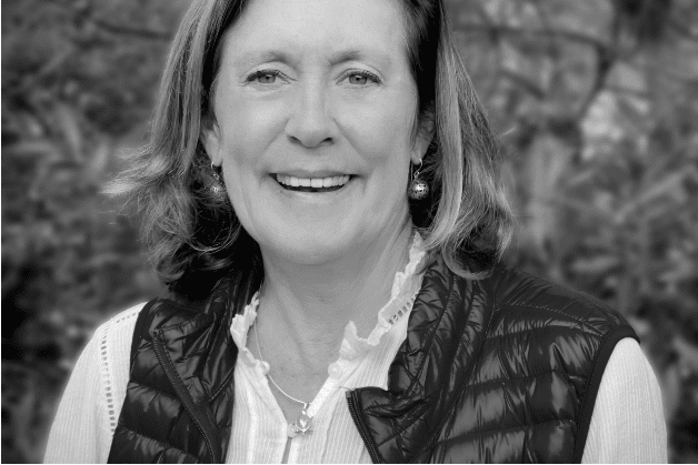 A woman smiling, wearing a white shirt and a dark puffy vest. The background is blurred with foliage. Black and white image.