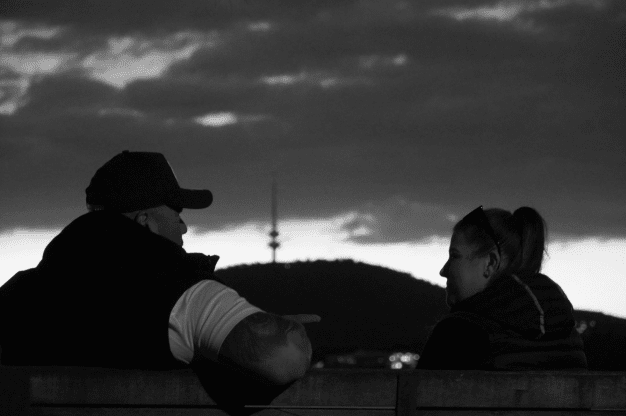 Two people sit on a bench, conversing against a backdrop of hills and a tower at dusk.