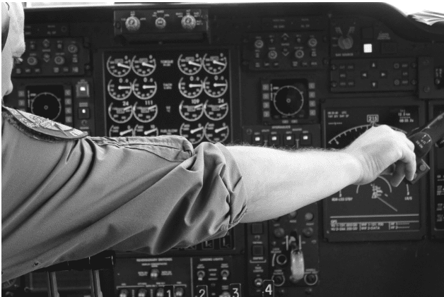 A pilot's arm reaching forward in an airplane cockpit, with various dials, gauges, and screens visible on the control panel.