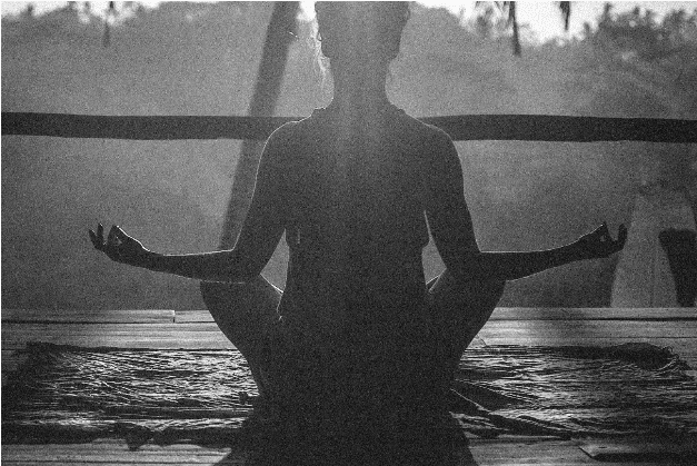 Silhouette of a person meditating in a seated position on a mat, with sunlight streaming from behind and a wooden structure in the background.