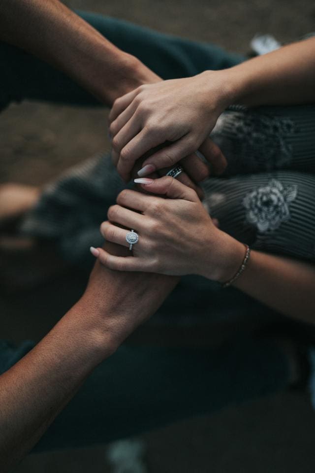 Two people gently holding hands, one wearing a ring with a circular setting, seated on the ground with floral-patterned clothing visible.