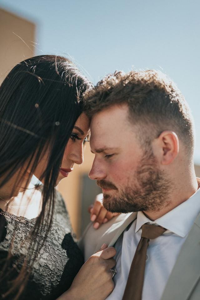 A man and woman in formal attire stand closely together outdoors, with the woman touching the man's shoulder and resting her forehead against his.