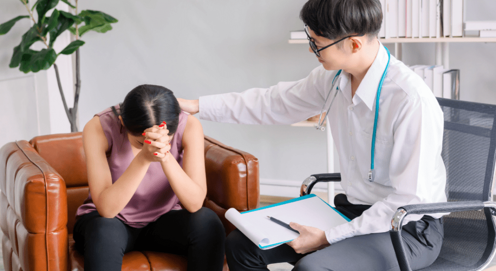 A person comforts another who is sitting with head down in a chair, holding a clipboard.