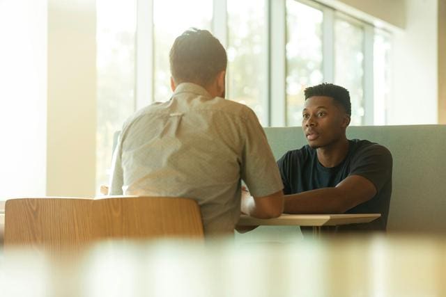 Two people sitting across each other at a table, engaged in a conversation in a sunlit room with large windows.