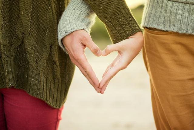 Two people form a heart shape with their hands, wearing green sweaters and standing closely together.