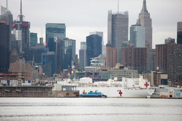 A hospital ship docked at a harbor with a city skyline in the background, featuring tall buildings and a cloudy sky.