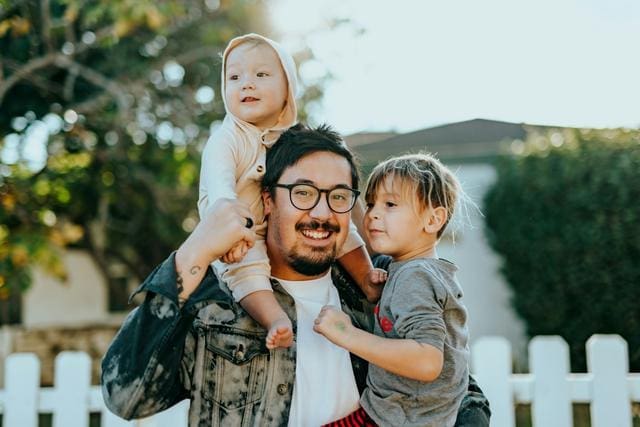 A person with glasses holds two young children, one on each arm, in front of a white fence.