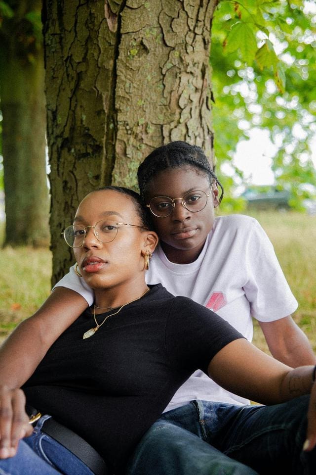 Two people are sitting outdoors against a tree, both wearing glasses and casual clothes, looking toward the camera.