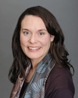 Adrienne W. Katzow, Ph.D. Smiling person with long brown hair, wearing a dark top and patterned scarf, against a gray background.