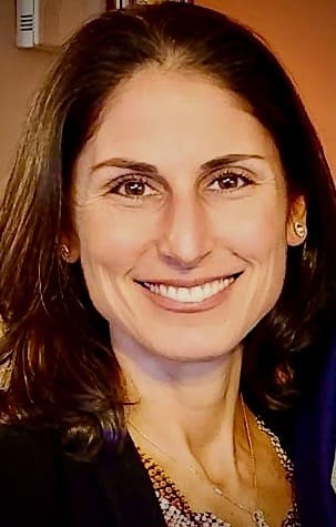 Smiling person with brown hair wearing a patterned top.