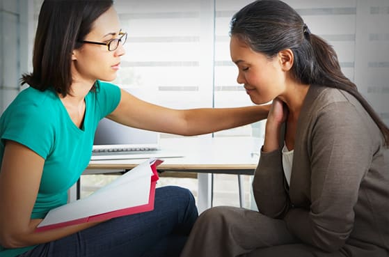 A woman in glasses comforts another seated woman by placing a hand on her shoulder in an office setting.