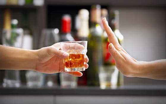 A hand offers a glass of whiskey while another hand gestures 'no' against a blurred background of bottles.