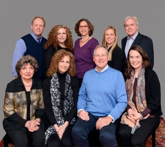 Nine people pose in two rows against a gray background, with four seated in front and five standing behind them.