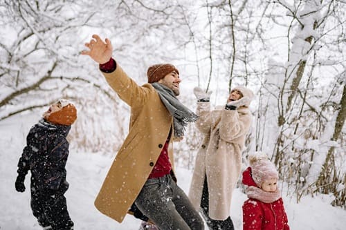 Four people, two adults and two children, are dressed in winter clothing and playing together in the snow among bare trees on a snowy day.