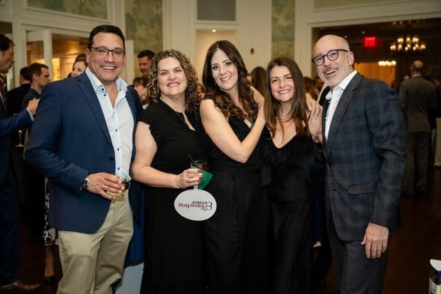 Five adults in formal attire stand together, smiling for a group photo at an indoor event or party. Other guests and chandeliers are visible in the background.