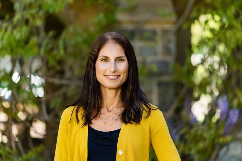Woman with long dark hair wearing a yellow cardigan and black top, standing outdoors in front of green foliage and stonework, smiling at the camera. Alexis