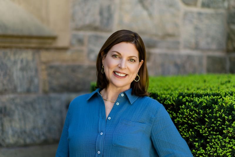A woman with shoulder-length brown hair in a blue button-up shirt stands outdoors, smiling, with a stone wall and manicured green hedge in the background Jen Jordan