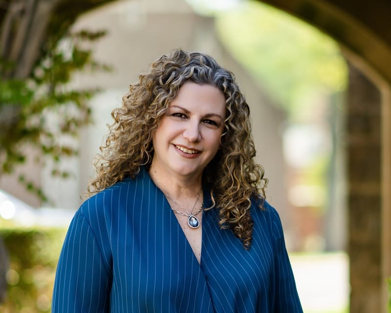 A woman with curly hair wearing a blue pinstripe blouse and necklace smiles outdoors under an archway. Jen Klein