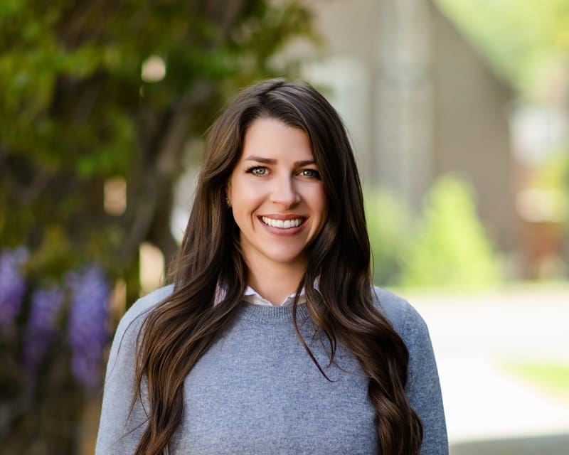 A woman with long brown hair, wearing a gray sweater over a collared shirt, smiles while standing outdoors with greenery and a blurred building in the background. Kara