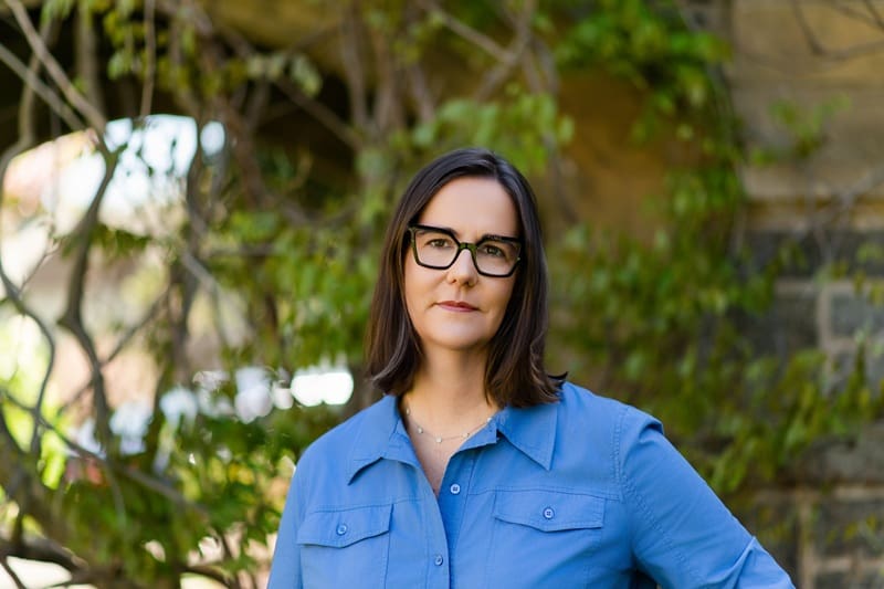 A woman with straight dark hair and glasses, wearing a blue button-up shirt, stands outdoors in front of greenery and a stone wall. Kathy