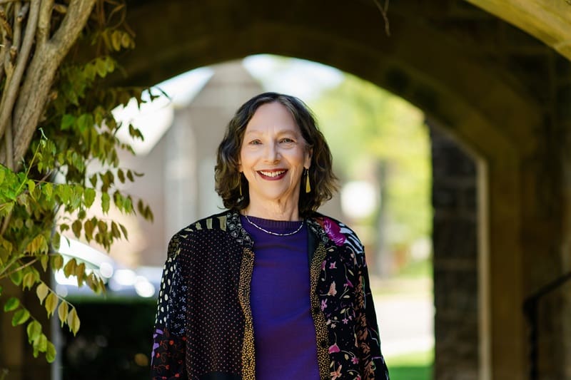 A woman with shoulder-length brown hair stands outdoors under an archway, smiling and wearing a patterned jacket over a purple top. Lynn