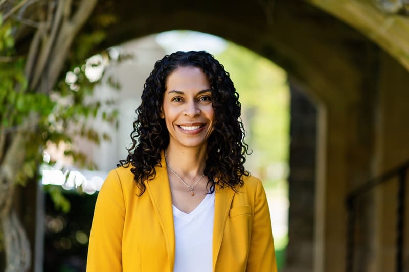 A woman with curly hair wearing a yellow blazer and white shirt smiles while standing outdoors under an archway. Natalie