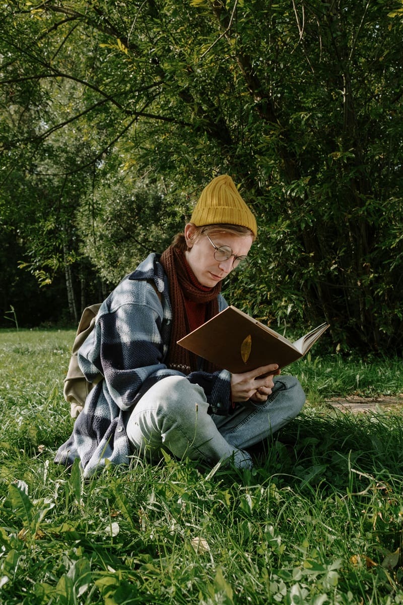 Person wearing a yellow beanie and glasses sits cross-legged on grass outdoors, reading a book, with trees and greenery in the background.