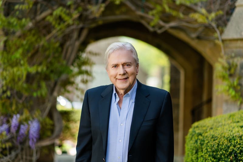 An older man with white hair in a dark suit and light blue shirt stands outdoors in front of a stone archway and greenery. Shoup