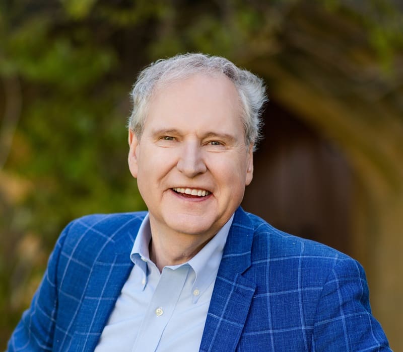 An older man with gray hair smiles, wearing a blue plaid blazer and light blue shirt, standing outdoors with blurred greenery and an arched doorway in the background. Virgil Roberson