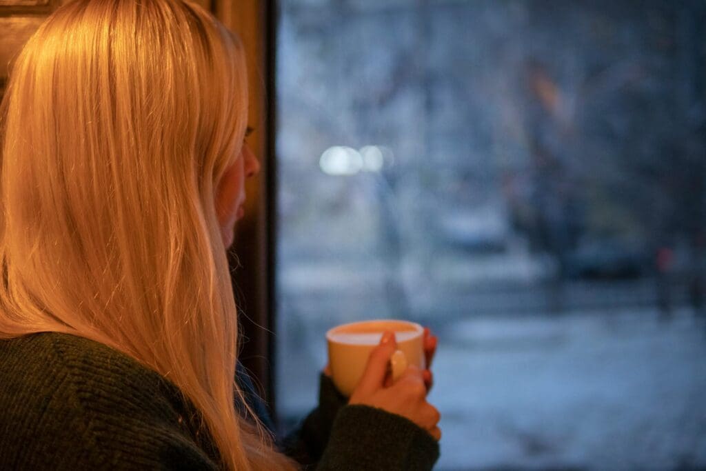 A person with long blond hair holds a cup and looks out of a window at a snowy, wintery scene outside.