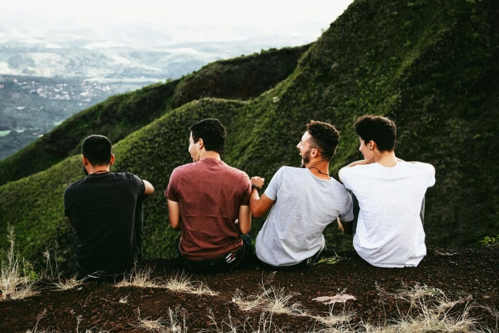 Four people sit on a grassy hillside facing away, overlooking a lush green valley and mountains in the distance.