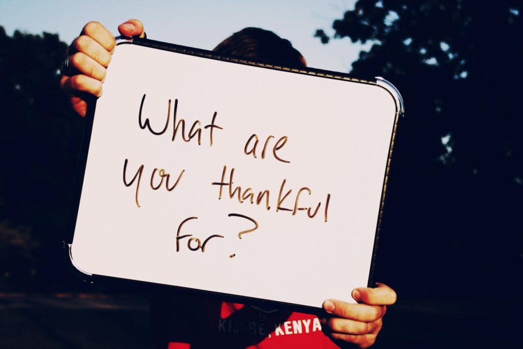 A person holds a whiteboard with the handwritten question “What are you thankful for?” outdoors, with trees visible in the background.