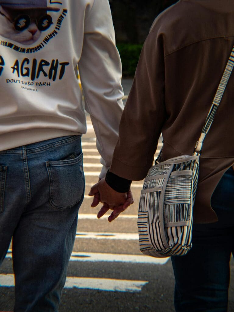 Two people holding hands while walking across a street, one wearing jeans and a white jacket, the other wearing a brown jacket and carrying a striped shoulder bag.