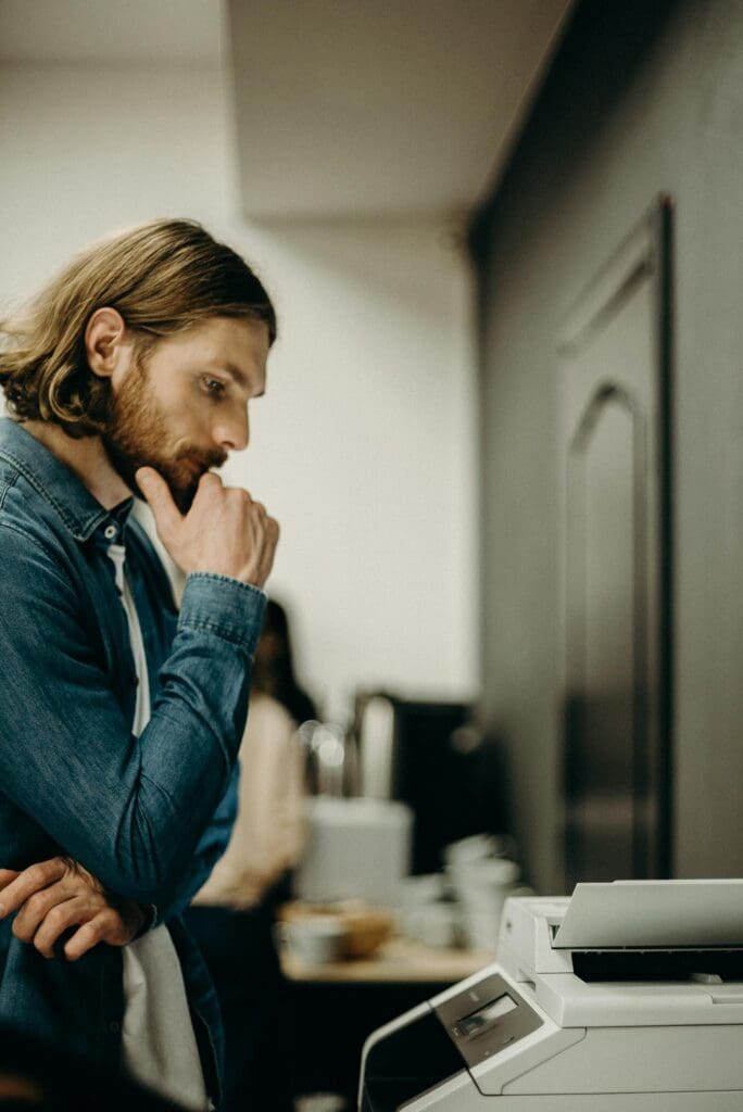 Man in a denim shirt stands in front of a printer, looking thoughtful with his hand on his chin.